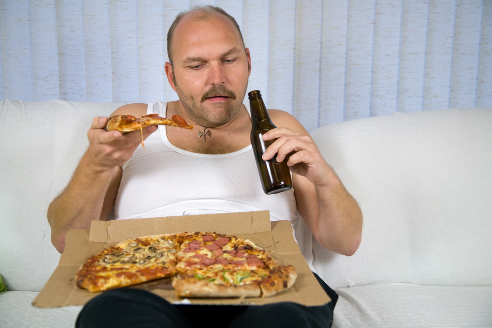 fat man sitting on couch drinking beer and eating pizza